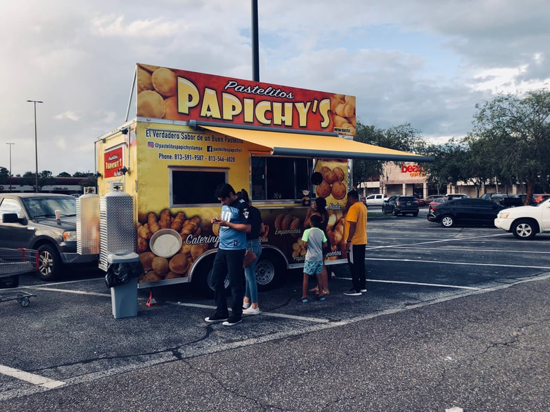 A food truck "Papichy's" in a parking lot surronded by people under an awning shading them